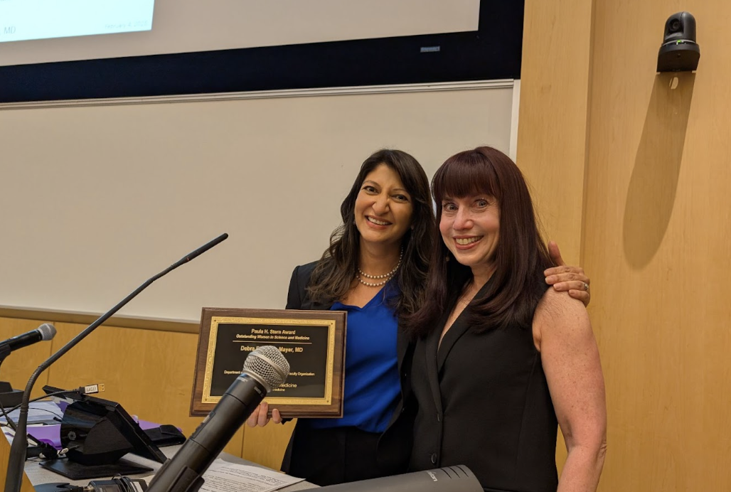 Two women pose behind a podium. One holds an award in her hand.